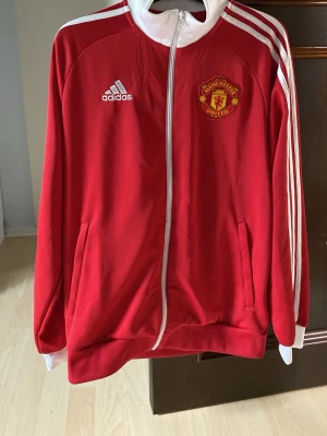 Röd Manchester United Tröja Adidas - Säljer en röd Manchester United tröja från Adidas med dragkedja, vita detaljer och klubbmärke på bröstet. Klassiska vita ränder längs ärmarna och två fickor framtill. Tillverkad i mjukt material, perfekt för dig som älskar fotboll och streetwear.