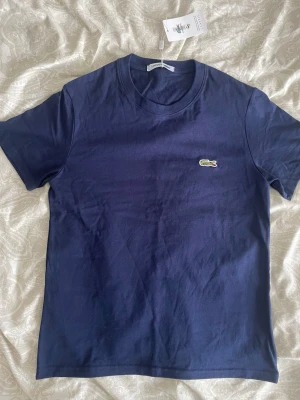 Marinblå t-shirt från Lacoste - Snygg marinblå t-shirt från Lacoste med klassisk krokodillogga på bröstet. T-shirten har rund halsringning och korta ärmar. Tillverkad i mjuk bomull för en skön känsla och stilren look.