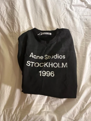 Svart långärmad Acne Studios t-shirt - Svart långärmad t-shirt från Acne Studios med vit text 'Acne Studios STOCKHOLM 1996' på bröstet. Klassisk rund halsringning och rak passform. Perfekt för dig som gillar stilrena och trendiga plagg.