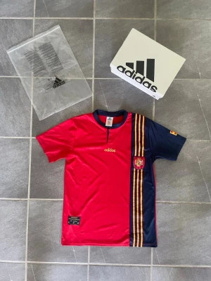 Adidas retro Spanien fotbollströja röd/blå - Säljer en officiell Spanien fotbollströja från Adidas i rött och marinblått med gula och röda ränder på sidan. Tröjan har korta ärmar, rund hals och Spanien-emblem på bröstet. Tillverkad i lätt och ventilerande material, perfekt för fotboll eller träning.