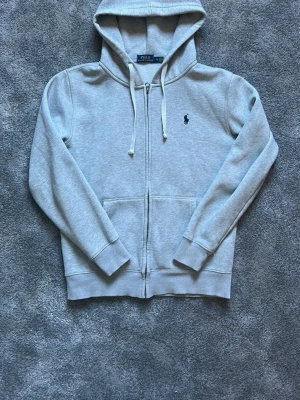 Polo Ralph Lauren hoodie - Snygg ljusgrå hoodie med dragkedja från Polo Ralph Lauren. Klassisk design med huva och vita dragsnören, två fickor framtill och den ikoniska lilla loggan broderad på bröstet. Priset är inte hugget i sten!