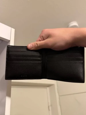 Svart plånbok från BOSS i läder - Snygg svart plånbok från BOSS i äkta läder med flera kortfack och RFID-skydd. Klassisk och stilren design med diskret BOSS-logga på insidan. Perfekt för att hålla ordning på kort och sedlar. Helt oanvänd i nyskick. OBS pris går att diskutera vid smidig affär. 