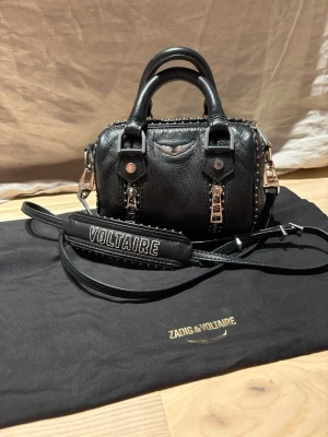 Svart handväska Zadig & Voltaire - Cool svart handväska i läder från Zadig & Voltaire med silverfärgade detaljer, dubbla handtag och avtagbar axelrem med broderad VOLTAIRE-text. Väskan har nitar längs kanterna och två dekorativa dragkedjor framtill. Perfekt för dig som gillar edgy och trendig stil. dustbag medföljer!🥰