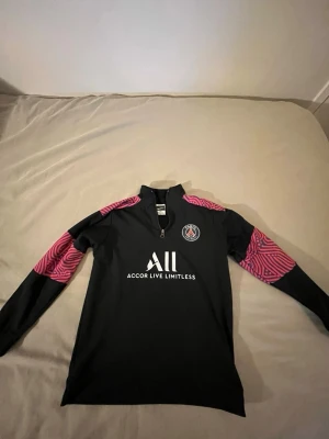 PSG träningströja svart Nike - Snygg långärmad träningströja från PSG i svart med rosa mönstrade detaljer på ärmarna. Halv dragkedja framtill och klubbmärke på bröstet. Tillverkad i ett lätt och stretchigt funktionsmaterial, perfekt för träning.