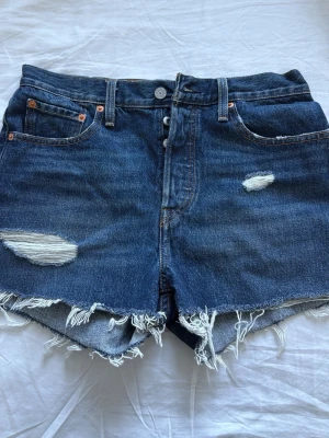 Blå denimshorts från Levi's - Säljer ett par klassiska blå denimshorts från Levi's med medel midja. Shortsen har slitna detaljer, fransig kant och femficksmodell. Perfekta för sommaren och ger en avslappnad vibe till din outfit.