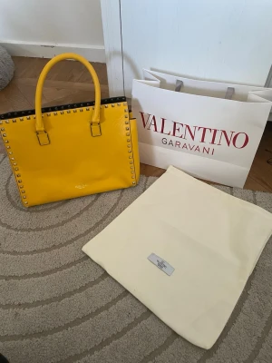 Gul Valentino Garavani handväska - Säljer en riktigt snygg gul handväska från Valentino Garavani. Väskan är i glansigt läder med silverfärgade nitar runt kanterna och har två rejäla handtag. Perfekt statement piece för dig som vill sticka ut! Dustbag och shoppingpåse från Valentino medföljer. Köptes på plick 5500kr. Fint skick utan defekter. Använd gärna köp nu💕