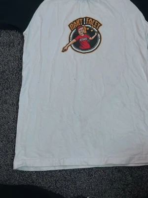 Vit t-shirt Rakt i Facet merch - Vit t-shirt med svart tryck på bröstet från Rakt i Facet. Klassisk passform med korta ärmar och rund hals. Trycket visar en tecknad figur med mikrofon och texten 'Rakt i Facet'. Perfekt för dig som gillar merch och vill sticka ut.