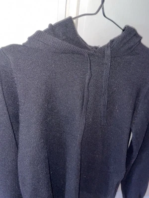 Snygg svart Kashmir hoddie med snygga snören  - En svart stickad hoodie med huva och dragsko. Tröjan har långa ärmar och ribbade muddar vid ärmslut och nederkant. Perfekt för en avslappnad och stilren look. Materialet känns mjukt och följsamt.