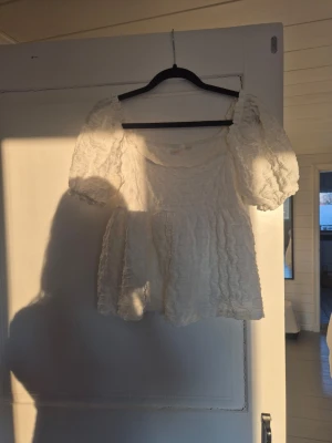 Vit puffärmad blus med struktur - Supersöt vit blus med puffiga korta ärmar och strukturerat tyg. Blusen har en luftig passform och en lätt rynkad effekt som ger en romantisk vibe. Perfekt till jeans eller kjol för en trendig look.