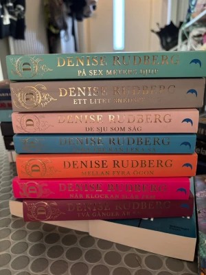 Bokserie av Denise Rudberg - Säljer en färgstark samling pocketböcker av Denise Rudberg. Omslagen är i olika färger som turkos, beige, rosa, orange och blå, med guldfolierad text och dekorativa detaljer. Perfekt för dig som gillar spännande och elegant skönlitteratur.