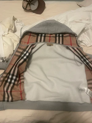 Grå hoodie från Burberry - Säljer en grå hoodie från Burberry med klassiskt rutigt foder på insidan av huvan och öppningen. Hoodien har dragkedja, ribbade muddar och är tillverkad i mjukt bomullsmaterial. Perfekt för dig som vill ha en stilren men ändå lyxig look.