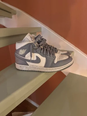 Gråvita Air Jordan 1 Mid sneakers - Snygga Air Jordan 1 Mid sneakers i grått och vitt läder med klassisk vinge-logga på sidan och perforerad tåbox. Skorna har grå snörning och vit sula, perfekt för dig som gillar streetwear och ikonisk design.