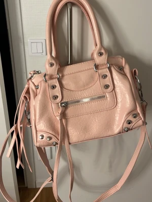 Ljusrosa handväska med nitar - Trendig ljusrosa handväska i konstläder med silverfärgade metalldetaljer, dragkedja framtill och dekorativa nitar. Väskan har både handtag och en lång axelrem samt långa tofsar för extra stilpoäng. Perfekt accessoar för att lyfta din outfit.