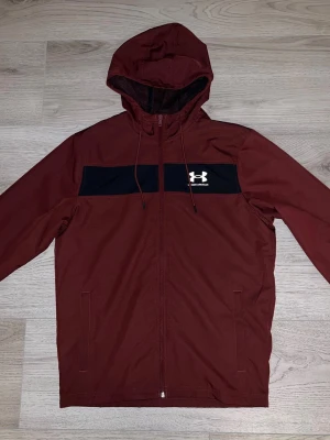Vinröd vindjacka från Under Armour - Snygg vinröd vindjacka från Under Armour med svart panel över bröstet och vitt logotryck. Jackan har huva med dragsko, hel dragkedja framtill och två sidofickor. Tillverkad i lätt och tåligt material, perfekt för blåsiga dagar.