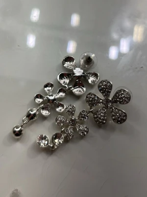Silvriga blommiga örhängen med stenar - Unika örhängen i silverfärg med flera blommor i olika storlekar. Varje blomma är dekorerad med gnistrande stenar som ger ett lyxigt intryck. Örhängena har en hängande design och är perfekta för dig som vill sticka ut med ett glittrande accessoar.