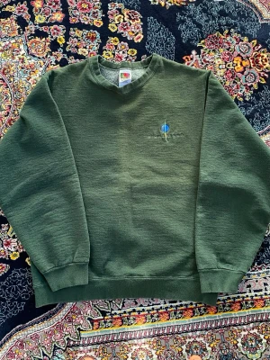 Grön sweatshirt från Fruit of the Loom - Mörkgrön sweatshirt från Fruit of the Loom med rund halsringning och diskret broderad detalj på bröstet. Tröjan har långa ärmar och ribbade muddar vid ärmslut och nederkant. Perfekt för en avslappnad och stilren look.