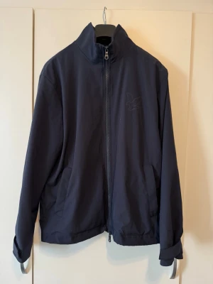 Mörkblå vindjacka från Lyle Scott  - Snygg mörkblå vindjacka från Ulle Scott med hög krage, hel dragkedja och två sidofickor. Jackan har minimalistisk design med diskret logga på bröstet och justerbara ärmslut. Perfekt för blåsiga dagar och enkel att matcha till olika outfits.
