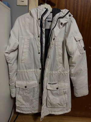 Everest Vinterjacka Parka storlek 34 - Snygg vit parkas från Everest med stor huva och flera praktiska fickor framtill. Jackan har dragkedja och tryckknappar, samt en ficka på ärmen. Perfekt för kalla dagar och har en sportig, clean look., Storlek 34