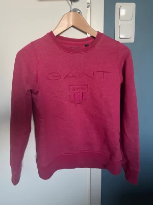 Rosa sweatshirt GANT - Säljer en mörkrosa sweatshirt från GANT med broderad logga och sköld på bröstet. Tröjan har rund halsringning, långa ärmar och ribbade muddar vid ärmslut och nederkant. Tillverkad i mjukt bomullsmaterial, perfekt för en avslappnad stil.