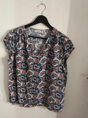 Mönstrad blus från flash Woman - Snygg blus från flash Woman med paisleymönster i blått, rött och brunt på vit botten. Blusen har V-ringning och korta ärmar. Tillverkad i ett lätt och luftigt material som passar perfekt till sommaren. Perfekt för dig som gillar färg och mönster. Skön och snygg. Normal passform och bra i storlek m,m. Storlek M. 