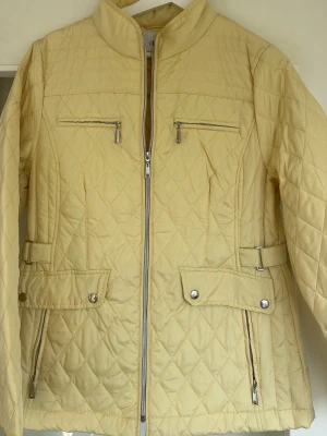 Ljusgul quiltad jacka från Isolde - Stilren ljusgul quiltad jacka från Isolde med dragkedja framtill, två bröstfickor med dragkedja och två sidofickor med lock och knapp. Jackan har justerbara spännen i midjan och ståkrage. Perfekt för dig som vill ha en snygg och praktisk jacka.