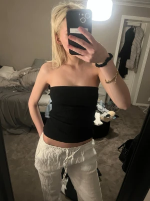 Svart ribbad bandeau tubtopp - Säljer en svart ribbad bandeau tubtopp som sitter tight och är perfekt för en enkel och clean look. Toppen är axelbandslös och har en croppad passform. Superenkel att matcha med olika stilar och accessoarer.