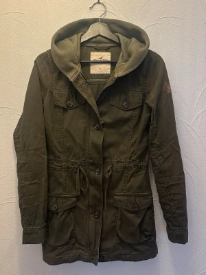 Hollister military jacket olive green hooded utility coat S - Hollister utility / military style jacket in olive green. Lightweight cotton with hood, adjustable waist and multiple pockets. Clean everyday piece with a fitted look.  Size S  Measurements (cm, laid flat): Waist 38 S2S 38 P2P 47 Length 73 Sleeve 65.5 Hips 50  Good used condition. Normal wear, no major flaws. See photos.