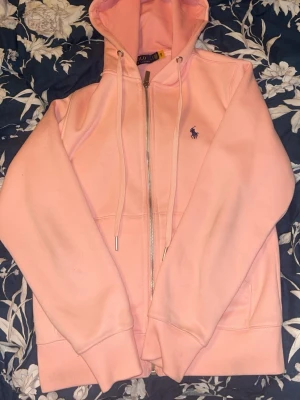 Ljusrosa hoodie från Ralph Lauren - Ljusrosa hoodie från Ralph Lauren med dragkedja framtill, justerbar huva med snören och den klassiska broderade loggan i mörkblått på bröstet. Tillverkad i mjukt bomullsmaterial och har fickor på sidorna. Perfekt för en avslappnad och stilren look.