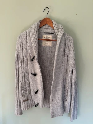 Ljusgrå stickad kofta från Blue Inc - Säljer en ljusgrå stickad kofta från Blue Inc med grov kabelstickning, bred sjalkrage och snygga träknappar. Koftan har långa ärmar, ribbade muddar och två fickor framtill. Perfekt för lager på lager och en chill look.