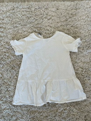 Vit blus med volang - Söt vit blus med diskret prickigt mönster, korta puffärmar och volang nedtill. Blusen har rund halsringning och en liten öppning bak i nacken. Har varit en lpngarmad tröja men har sytt den. Har två små hål på sidorna.