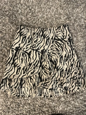 Zebramönstrad kortkjol med volang - Snygg kortkjol med zebramönster i svart och vitt. Kjolen har en liten volang längst ner som ger extra attityd. Perfekt för dig som vill sticka ut med ett djuriskt print. Resår i midjan för skön passform.