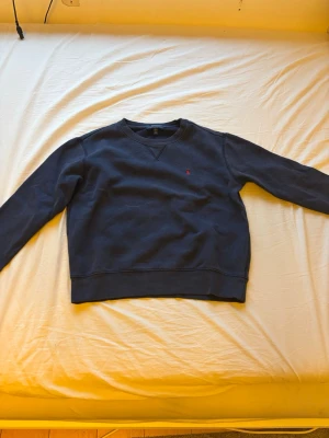 Mörkblå sweatshirt från Ralph Lauren - Klassisk mörkblå sweatshirt från Ralph Lauren med rund halsringning och diskret röd logga broderad på bröstet. Tröjan har ribbade muddar vid ärmslut och nederkant. Perfekt för en avslappnad och stilren look.