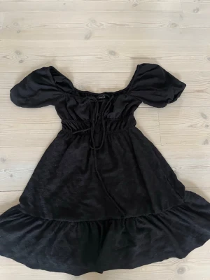 Svart kort klänning med puffärm - Snygg svart kort klänning med puffärmar och snörning i midjan. Klänningen har en lätt volang nedtill och en mjuk, något glansig yta. Perfekt för dig som gillar en feminin och trendig stil.