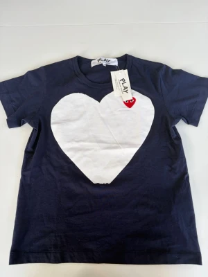 Mörkblå Comme des Garçons PLAY t-shirt - Säljer en mörkblå t-shirt från Comme des Garçons PLAY med ett stort vitt hjärta på bröstet. Klassisk rund hals och korta ärmar. Tillverkad i mjuk bomull, perfekt för en clean och trendig look.