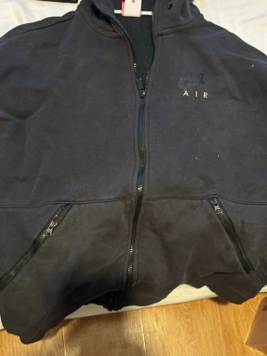 Svart Nike Air hoodie med dragkedja - Svart hoodie från Nike Air med hel dragkedja framtill och två snedställda fickor med dragkedja. Klassisk huva och diskret Nike Air-logga på bröstet. Tillverkad i mjukt sweatshirtmaterial som är skönt att bära.