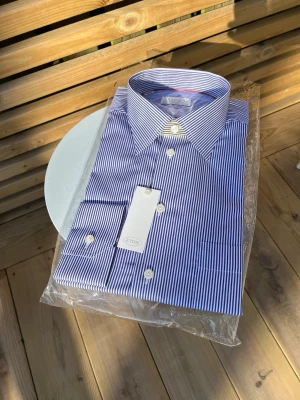 Blårandig skjorta från Eton - Klassisk blårandig skjorta från Eton med vit knappstängning och bred krage. Skjortan är tillverkad i bomull och har en tidlös design med smala vertikala ränder. Perfekt för dig som gillar stilrena och eleganta plagg.Aldrig använd, prislapp finns med Påsen man får till är även med. Ställ gärna frågor. Nu pris 1700
