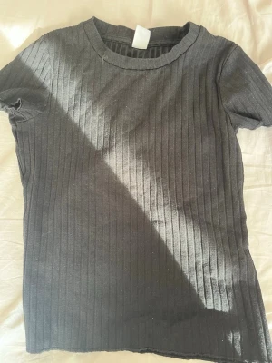 Svart ribbad topp med kort ärm - En svart ribbad topp med korta ärmar och rund hals. Toppen har en slim passform och är tillverkad i ett mjukt, stretchigt material som sitter snyggt på kroppen. Perfekt att styla med jeans eller kjol för en clean look.