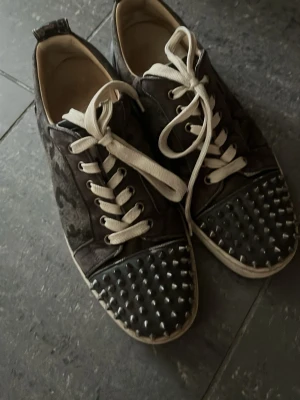 Svarta sneakers med nitar - Unika svarta sneakers från Christian Louboutin med vita snören och coola silverfärgade nitar på tån. Skorna har en låg profil och är tillverkade i en mix av mocka och läder. Perfekta för dig som vill sticka ut med din stil.