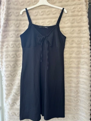 Svart ärmlös klänning med knyt - Svart långklänning med smala axelband och v-ringning. Klänningen har en snygg knytning framtill och är tillverkad i ett mjukt, lätt material som passar perfekt till varma dagar. Enkel och stilren design som är lätt att matcha.