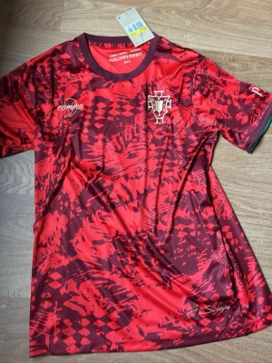 Portugal Nike röd matchtröja - Snygg Portugal matchtröja från Nike i rött med mörkröda och svarta mönster. Tröjan har korta ärmar, rund hals och Portugal-emblem på bröstet. Tillverkad i lätt och ventilerande material, perfekt för fotboll eller träning. Tröjan kan bytas mot parfym