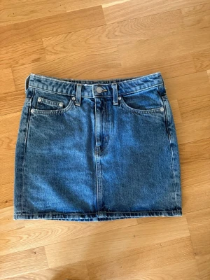 Blå jeanskjol från Weekday - Säljer en klassisk blå jeanskjol från Weekday med fem fickor, bälteshällor och knappstängning fram. Kjolen har en rak passform och är i ett tidlöst denimtyg som passar till allt. Perfekt basplagg för garderoben! Aldrig använd