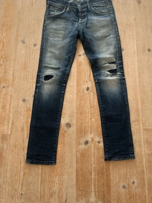 Blå slitna jeans med hål - Säljer ett par blå jeans med slitningar och hål på båda benen. Jeansen har en klassisk femficksmodell, normal midja och smal passform. Färgen är tvättad blå med ljusare partier och tydliga slitningar för en trendig, avslappnad look.
