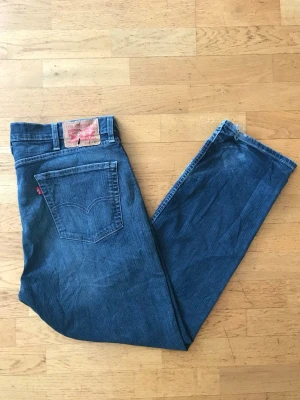 Levi's blå raka jeans 505 - Klassiska blå Levi's 505 jeans med rak passform och fem fickor. Jeansen har den ikoniska röda Levi's-lappen på bakfickan och patch i läder bak i midjan. Tillverkade i slitstark denim, perfekta för en avslappnad och tidlös stil.