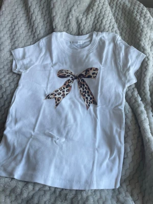 Vit t-shirt med leopardrosett - Snygg vit t-shirt med ett tryck av en rosett i leopardmönster. Klassisk rund halsringning och korta ärmar. Perfekt för dig som vill ha en basic tee med en twist. Sömmen har gått upp lite men är enkelt att laga. Skriv gärna om funderingar 💕