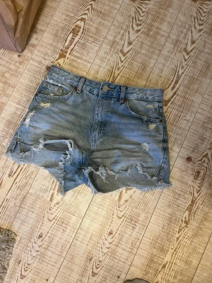 Blåa high waist jeansshorts - Säljer ett par blåa high waist jeansshorts med råa kanter och slitna detaljer. Shortsen har fem fickor och knappgylf. Storlek 36