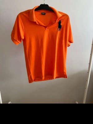 Orange pikétröja från Ralph Lauren - Snygg orange pikétröja från Ralph Lauren med klassisk krage och korta ärmar. Stor logga i mörkblått på bröstet och siffror på ena ärmen. Tillverkad i mjuk bomull som känns skön mot huden. Perfekt för en avslappnad men stilren look.