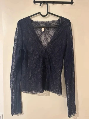 Mörkblå spetsblus med knyt - Säljer en mörkblå, transparent spetsblus med långa ärmar och v-ringning. Blusen har ett snyggt knyt framtill och är figurnära i modellen. Perfekt att styla över ett linne eller topp för en trendig look.