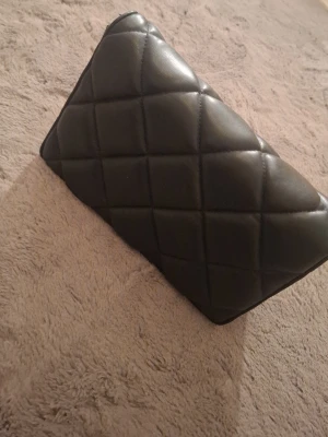 Svart quiltad kuvertväska - Stilren svart kuvertväska i quiltat konstläder med rutmönster. Väskan har en klassisk rektangulär form och känns både trendig och tidlös. Perfekt  när du vill addera en elegant touch till din outfit.