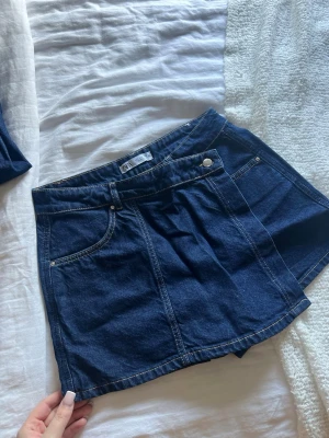 Mörkblå jeansskort från ZARA - Snygg mörkblå jeansskort från ZARA med klassiska fickor fram och knappdetalj i midjan. Skorten har en rak passform och är gjord i denim med kontrastsömmar. Perfekt för en trendig och avslappnad look.