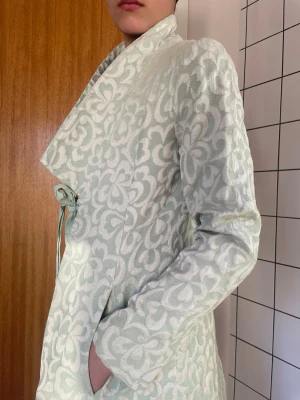 Ljusgrön mönstrad kappa Noa Noa - Bröllop, representativt på jobbet, bal, student eller fest. En väldigt fin kappa -Miss Vintage - by Noa Noa med blommigt mönster i vitt. Kappan har knytband i midjan, stora slag och fickor. Materialet är vävt och ger en exklusiv känsla.